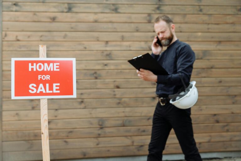 Real estate agent on phone with 'Home for Sale' sign outside a property.