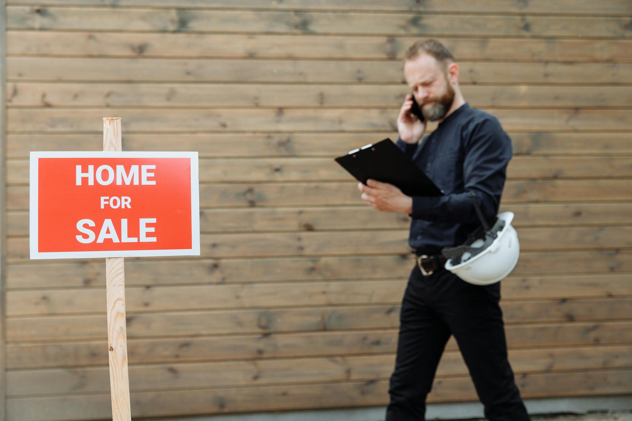 Real estate agent on phone with 'Home for Sale' sign outside a property.
